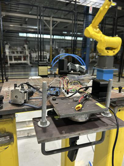 Photo of the built test rig on a steel table with a stepper motor, Arduino, and load cell, with an industrial robot behind it